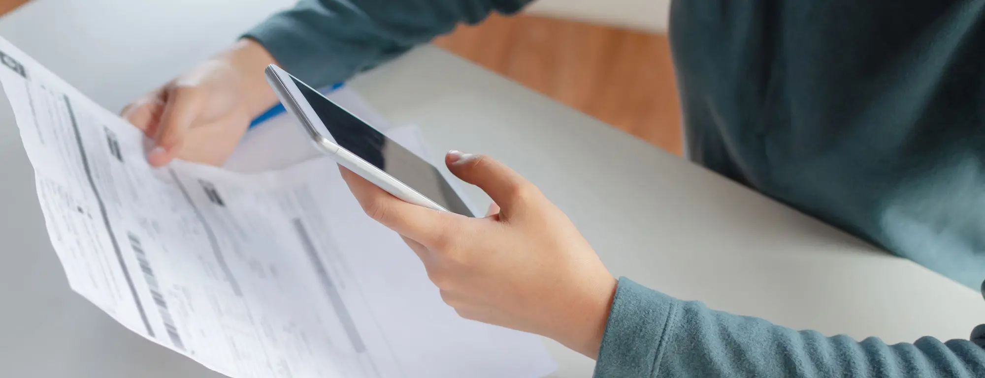 A person holds a smartphone in their right hand while reviewing documents with their left hand on a table.